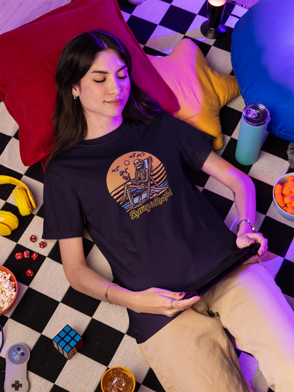Person wearing a black t-shirt with a graphic design, lying on a checkered floor with colorful pillows and snacks. T-shirt image is a skeleton on a beach chair, drinking a cocktail, with the tagline “Rotting is Restful.” 
